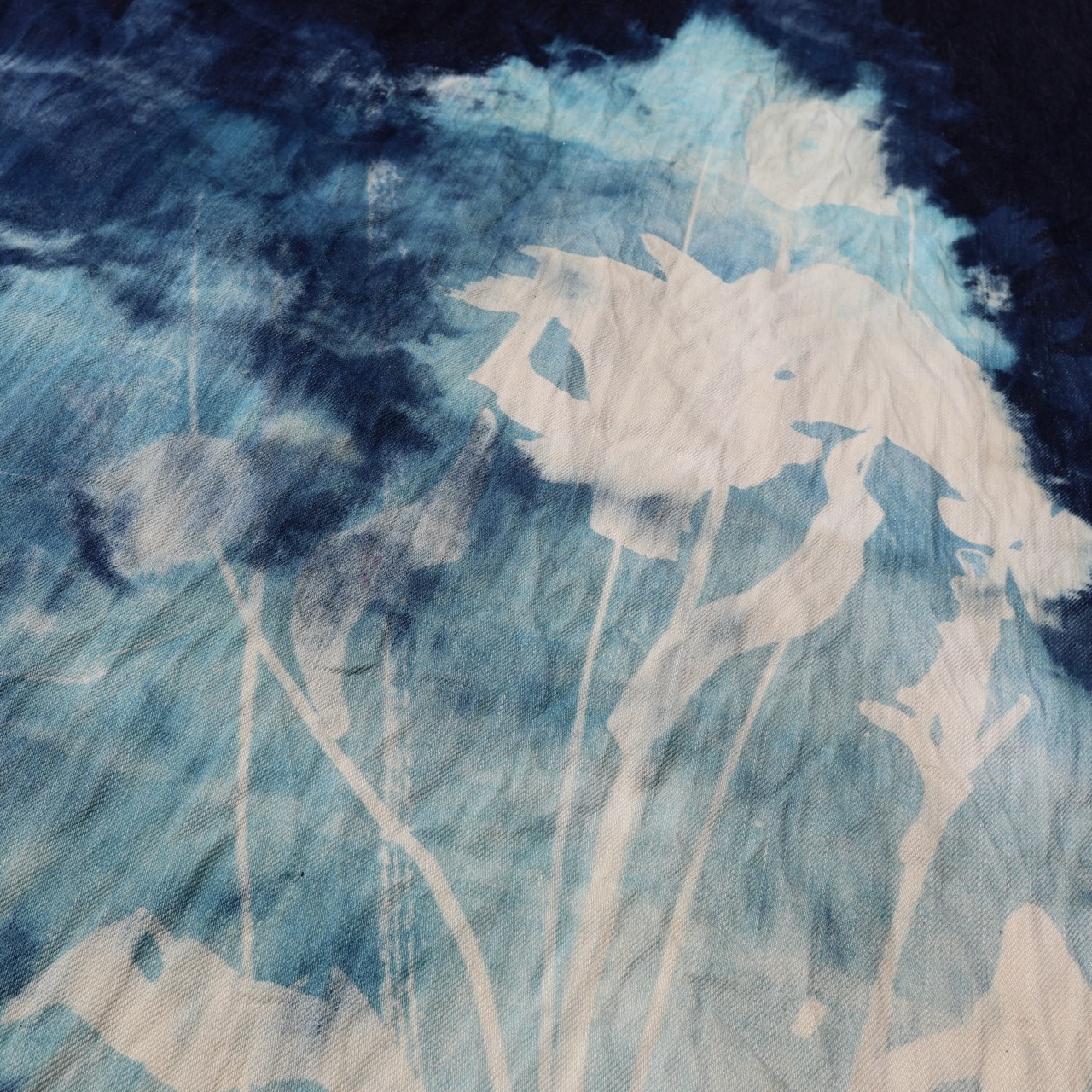 An abstract textile pattern in shades of blue and white, featuring silhouettes of flowers and stems. The design displays a blend of soft textures and colors, creating a dynamic visual interest.