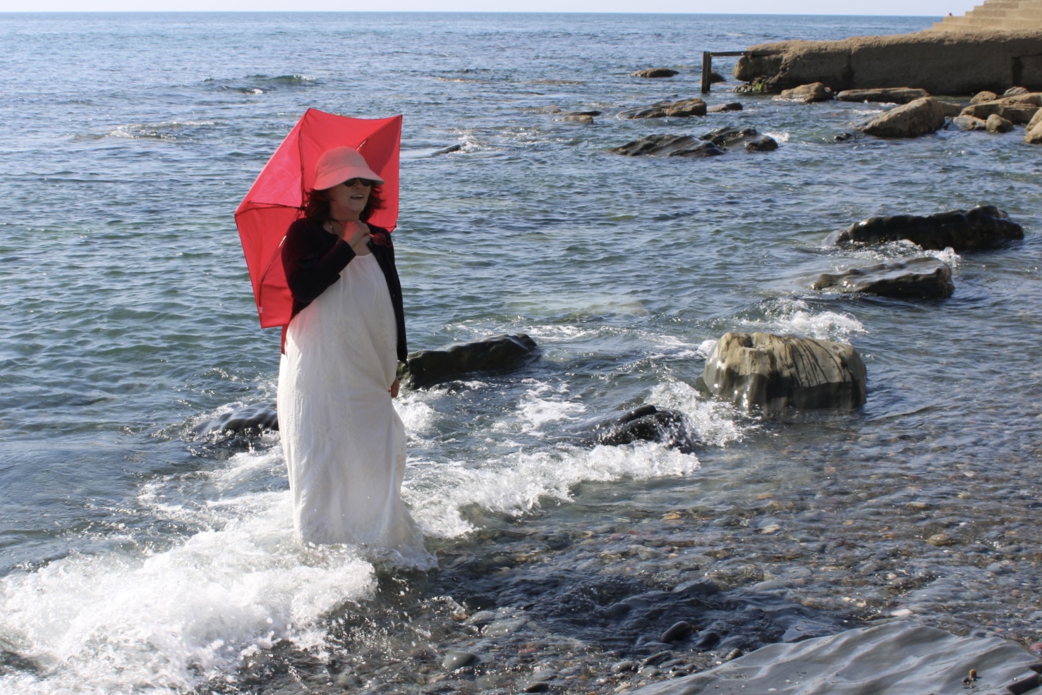 A person in a white dress stands in shallow seawater holding a bright red umbrella, with rocks and a distant shoreline in the background.