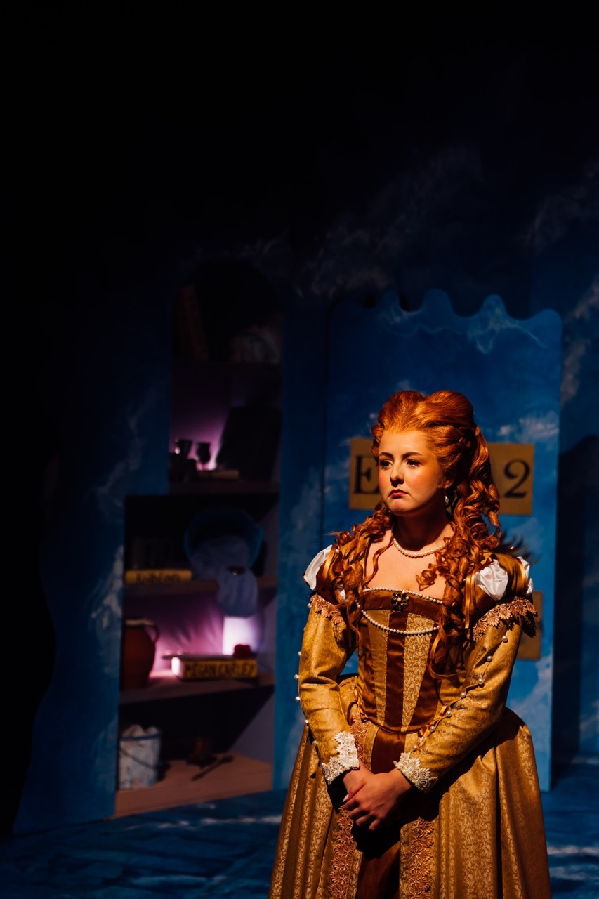 Woman in golden period dress with curled hair on dimly lit theatre stage with blue backdrop and shadowy set pieces.
