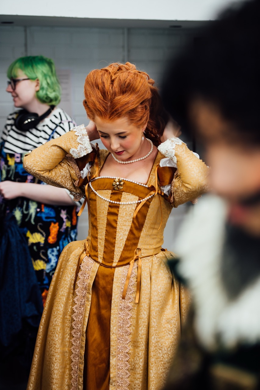 Woman in elaborate gold period dress with lace trim and pearl necklace adjusting her ornate red hair. Person with green hair visible in background.