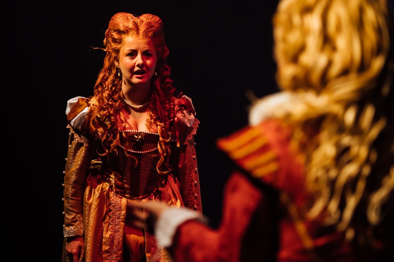 Woman in elaborate red period costume with long auburn hair performing on stage, facing another performer in foreground.