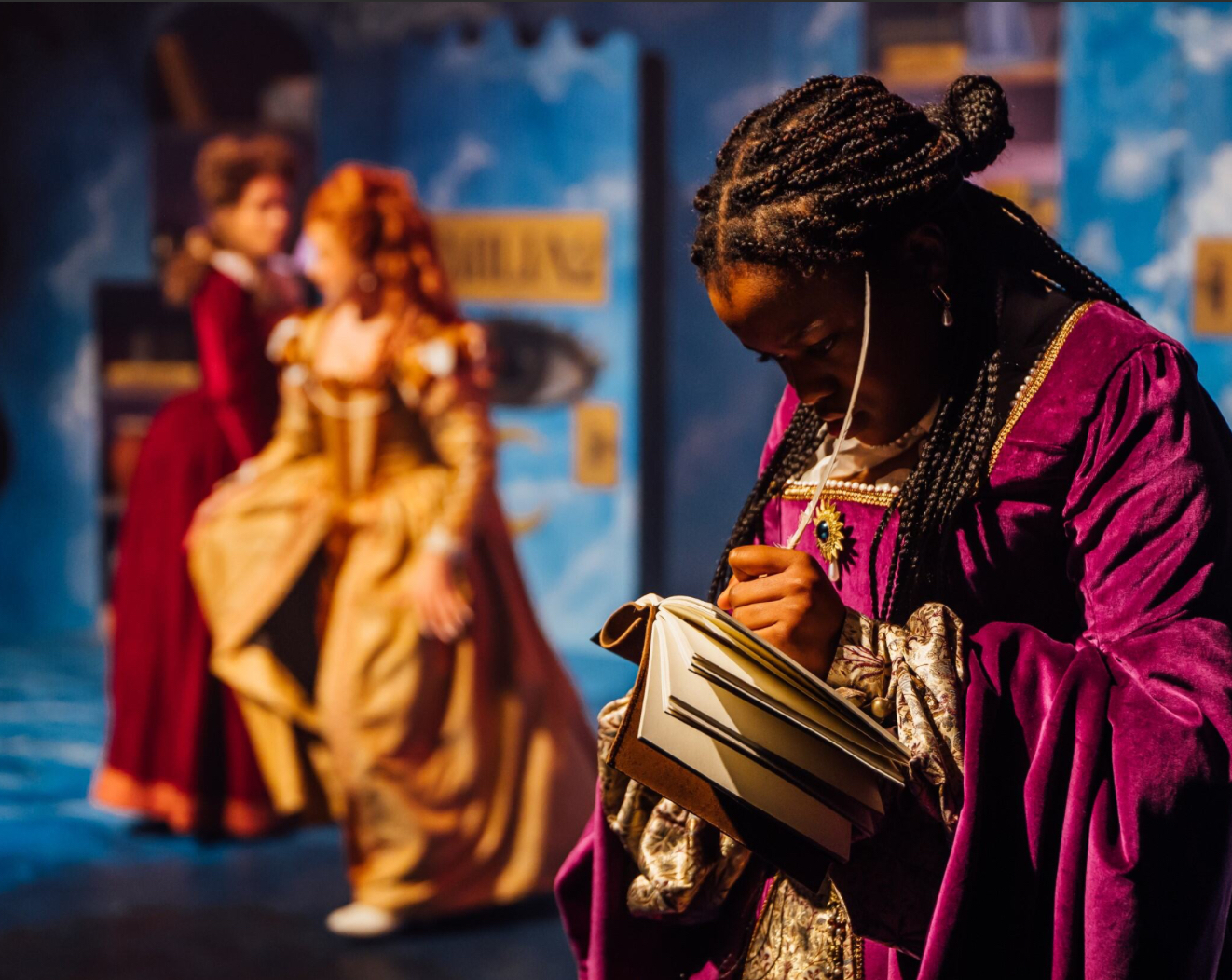 A BA (Hons) Acting student dressed in a pink 16th century gown writes in a book on stage in "Emilia!"