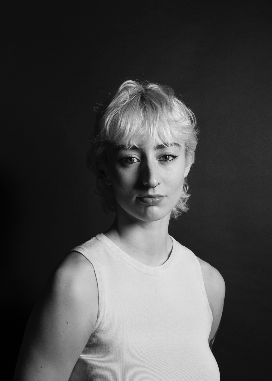 Black and white portrait of person with short blonde hair wearing white sleeveless top against dark background.