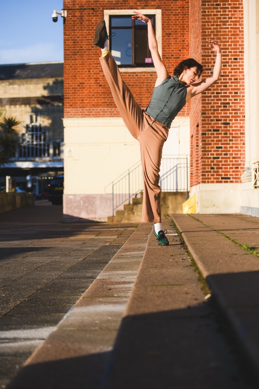 Person performing high kick stretch on paved courtyard against red brick building with white stairs and railings.