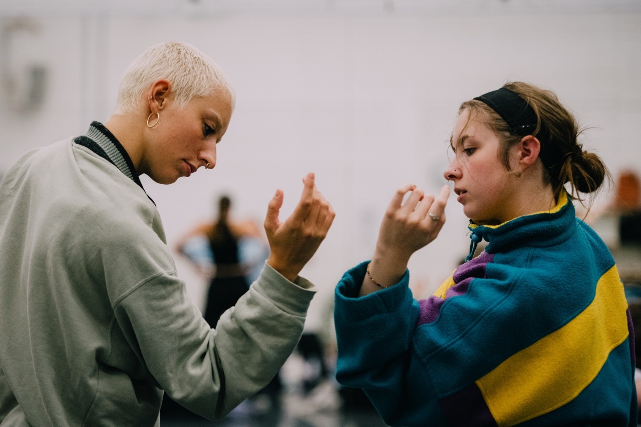 Two women face each other with raised hands in defensive positions. Left wears grey hoodie, right wears blue and yellow top with hair in bun.