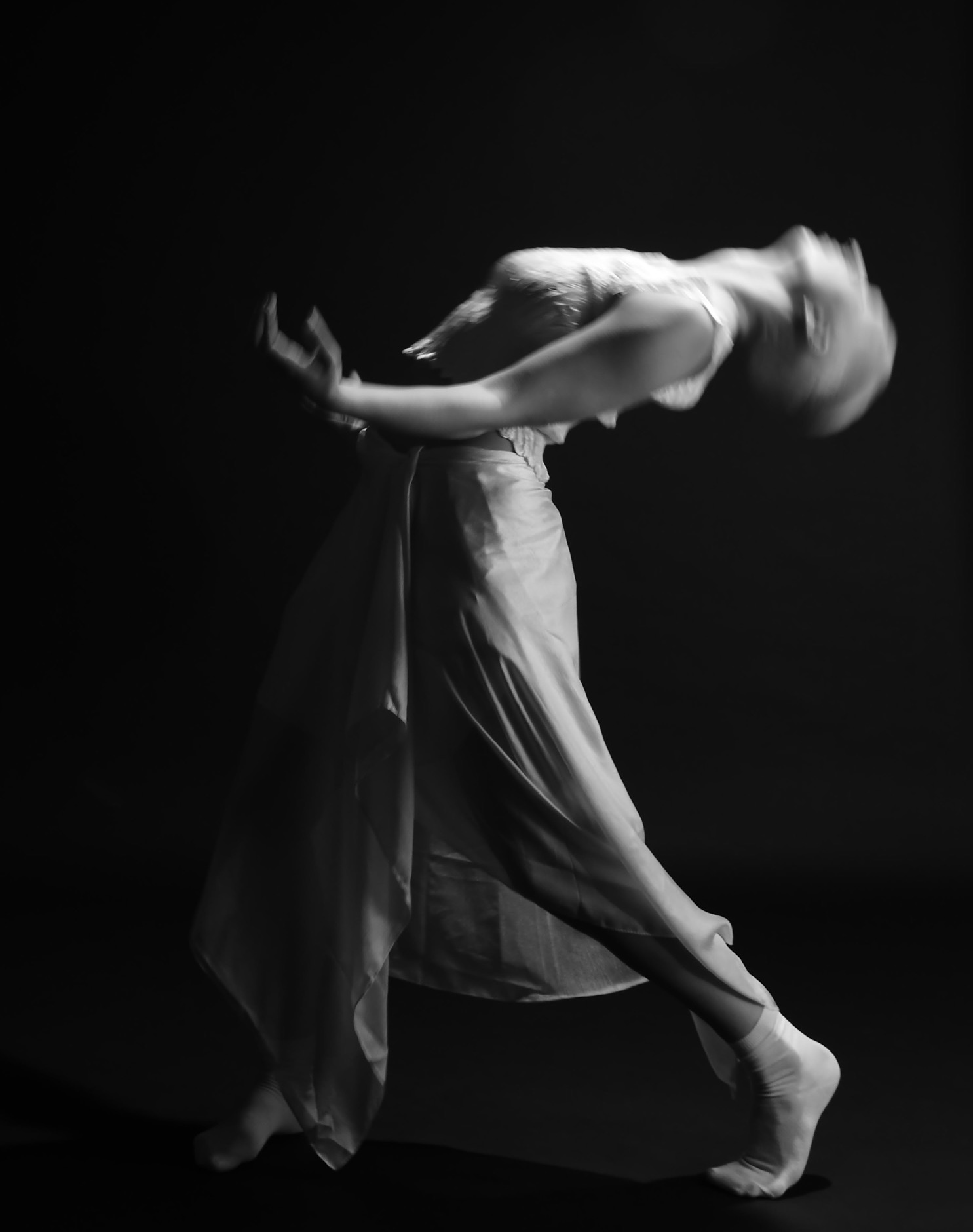 Black and white image of dancer in flowing skirt, arms extended horizontally with motion blur, against dark background.