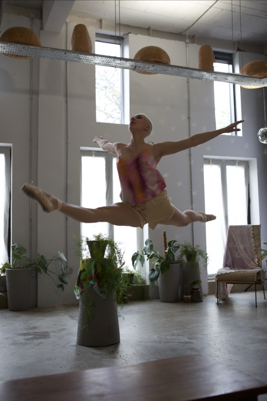 Dancer performing split leap in bright studio with large windows, plants, and pendant lights hanging from ceiling.