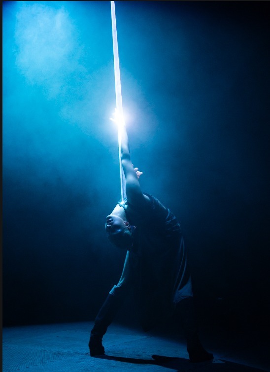 Person in dark clothing under illuminated ray of blue light overhead against blue-lit background with bright white light source above.