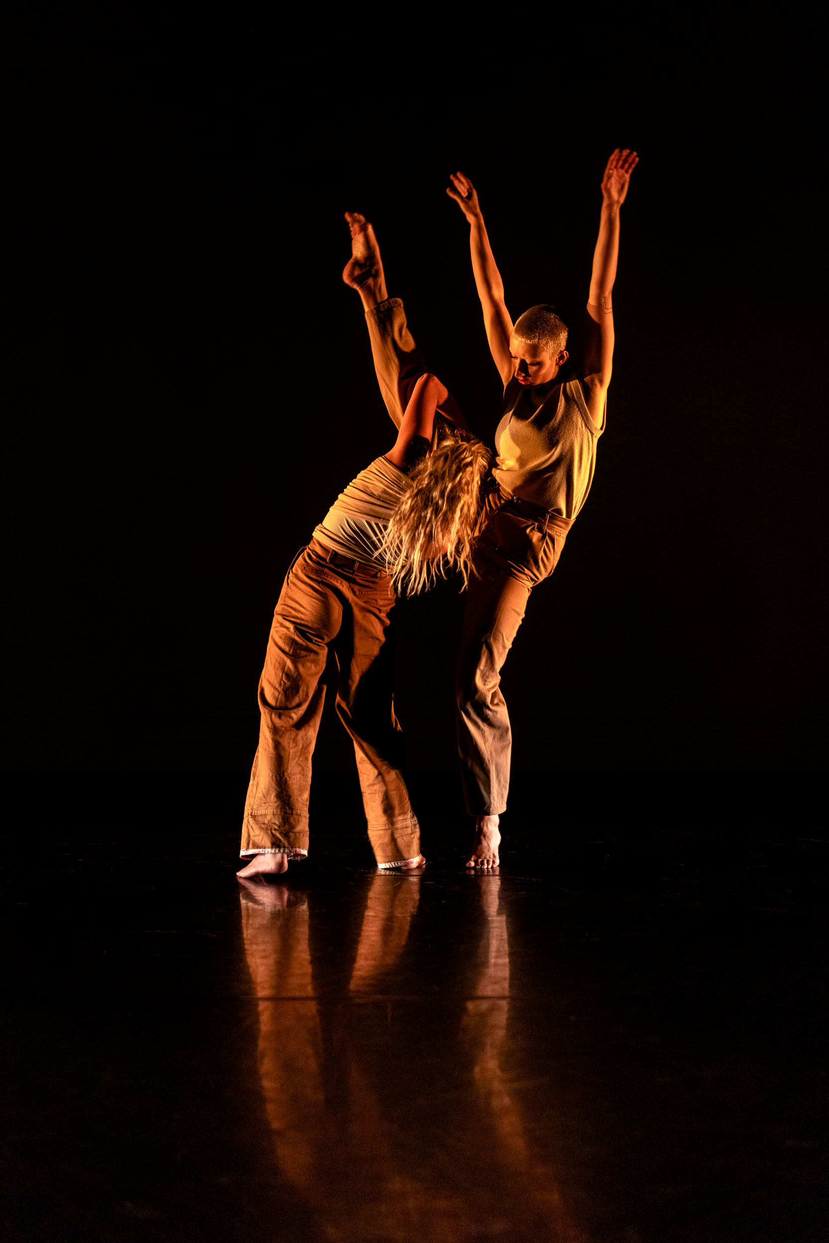Two dancers performing on stage with arms raised, illuminated by warm orange lighting against black background on reflective floor.