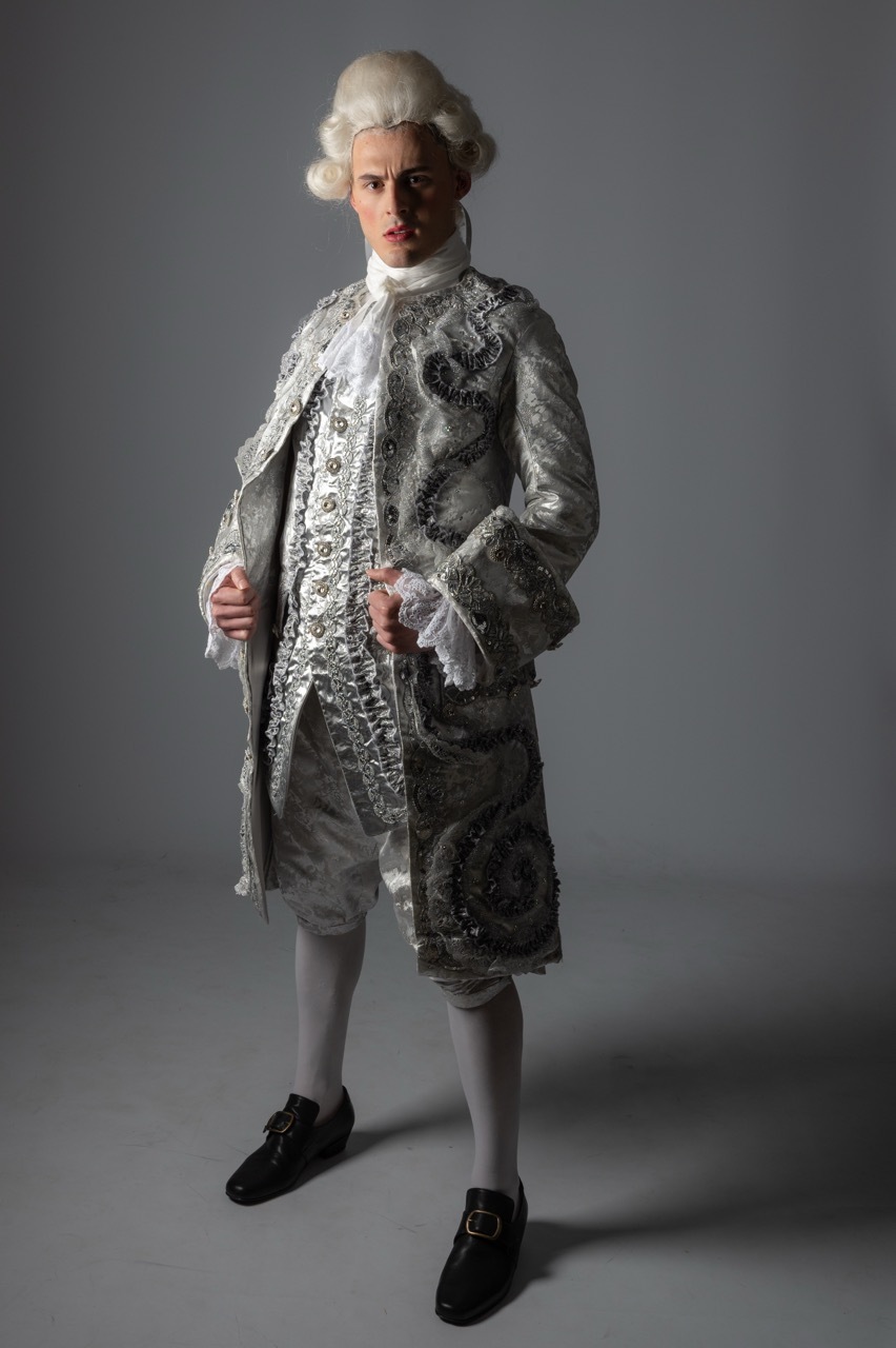 Man in 18th-century costume with powdered wig, ornate silver brocade coat, knee breeches, white stockings, and black buckled shoes.