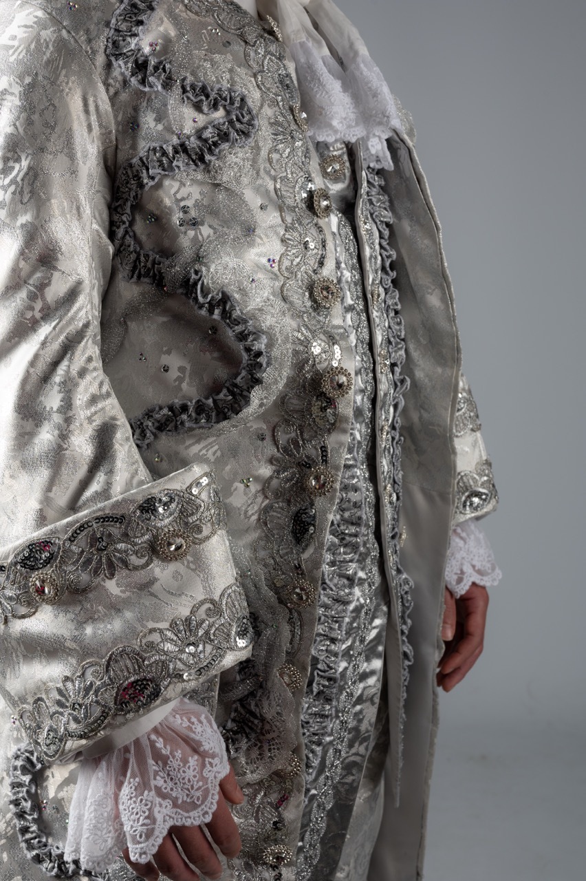 Grey translucent coat with ornate metallic embroidery, beading, and lace trim worn over layered garments against neutral background.