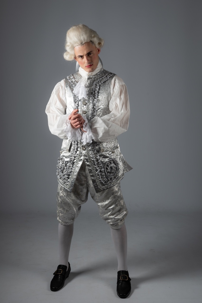 Man in 18th-century costume with white powdered wig, ornate silver waistcoat, white shirt, knee-length breeches, and black shoes.