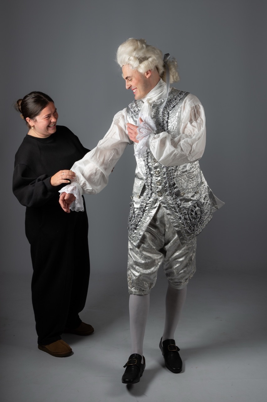 Man in 18th-century silver brocade waistcoat, white shirt, powdered wig shaking hands with woman in black modern clothing.