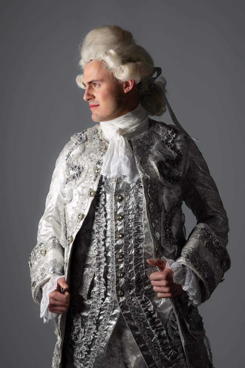 Man in 18th-century costume with white powdered wig, ornate silver brocade coat, white ruffled shirt, and black ribbon tie.