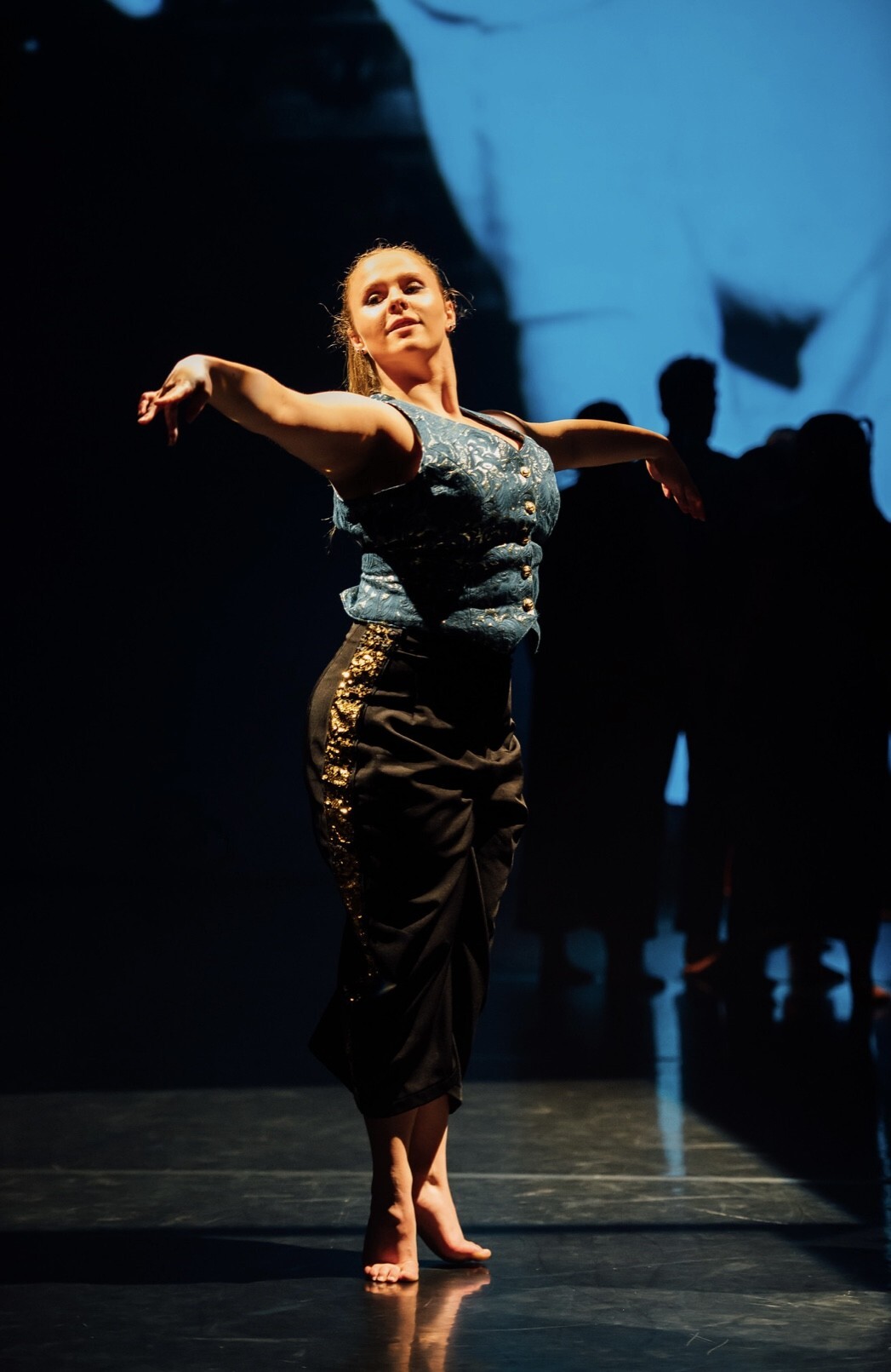 Woman dancing on stage in light blue top and dark skirt, arms extended, with shadowy figures and blue lighting in background.