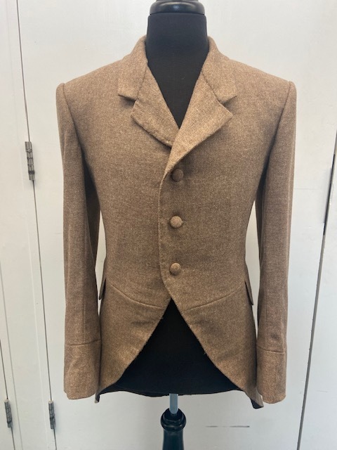 Beige tailored jacket with three buttons displayed on black mannequin torso against white background with glass cabinet doors visible.
