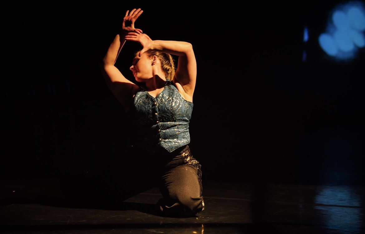 Woman kneeling on stage with arms raised above head, wearing grey top and brown skirt, illuminated by warm stage lighting.