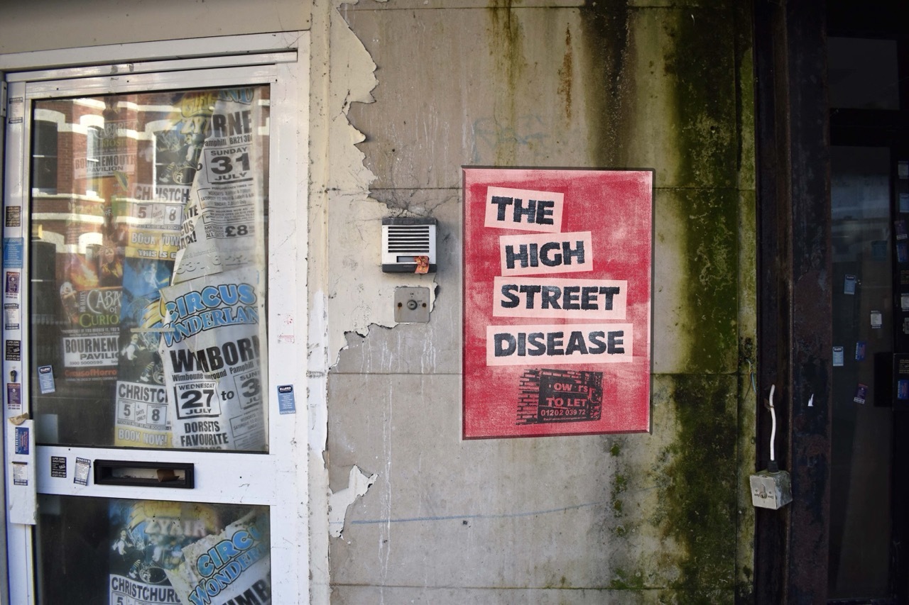 Red poster on a derelict wall with black text that reads: 'The High Street Disease'.