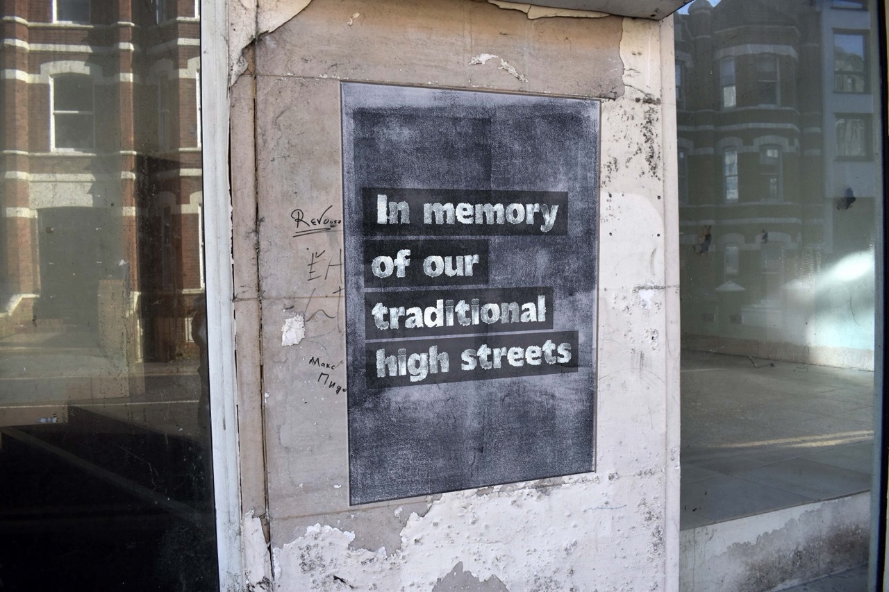 Grey poster with text that reads 'In memory of our traditional high streets' on an abandoned store front.