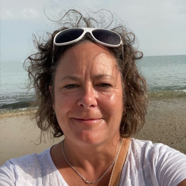 Woman with curly hair and white sunglasses on her head, wearing white top and gold necklace, seaside background with waves.