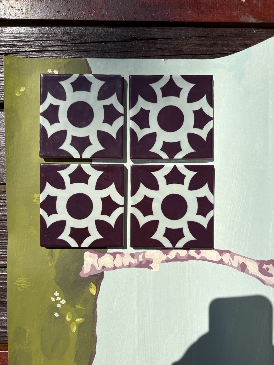 Four square tiles arranged in a two-by-two grid, featuring a design of alternating purple circles and floral shapes on a light background. The tiles are placed on a painted surface with hints of green and a abstract decorative element.
