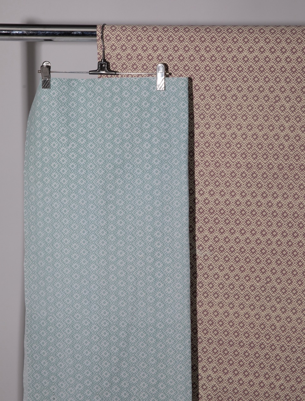 Two pieces of fabric hanging on a display rack. The left fabric is a pale blue with a small, repeating pattern, while the right fabric is a warm brown with a similar pattern in darker shades. Both are clipped at the top.