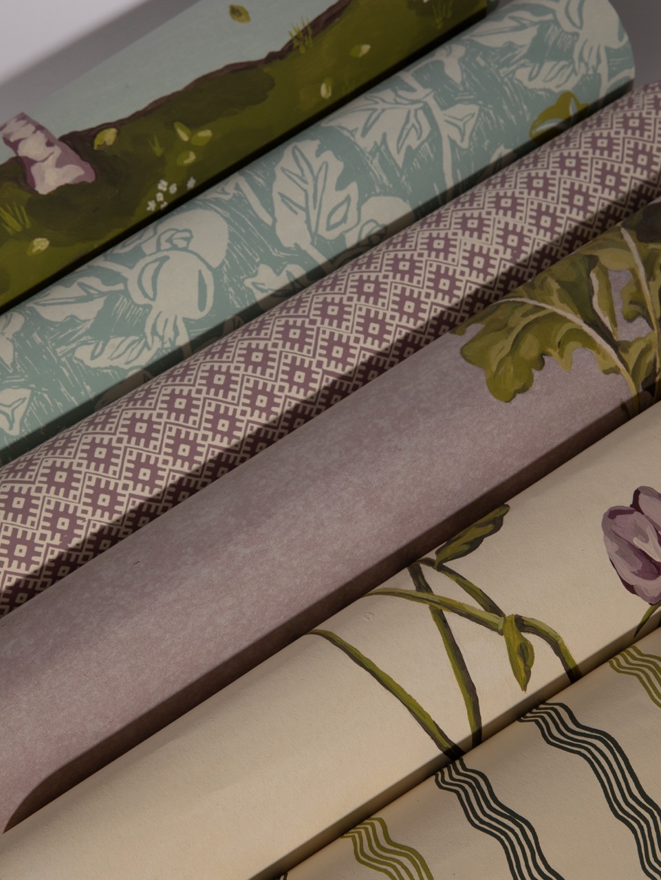 Rolls of wallpaper featuring various patterns, including floral designs, geometric shapes, and botanical motifs. The wallpaper rolls are stacked, displaying a mix of colours such as green, brown, mauve, and cream.