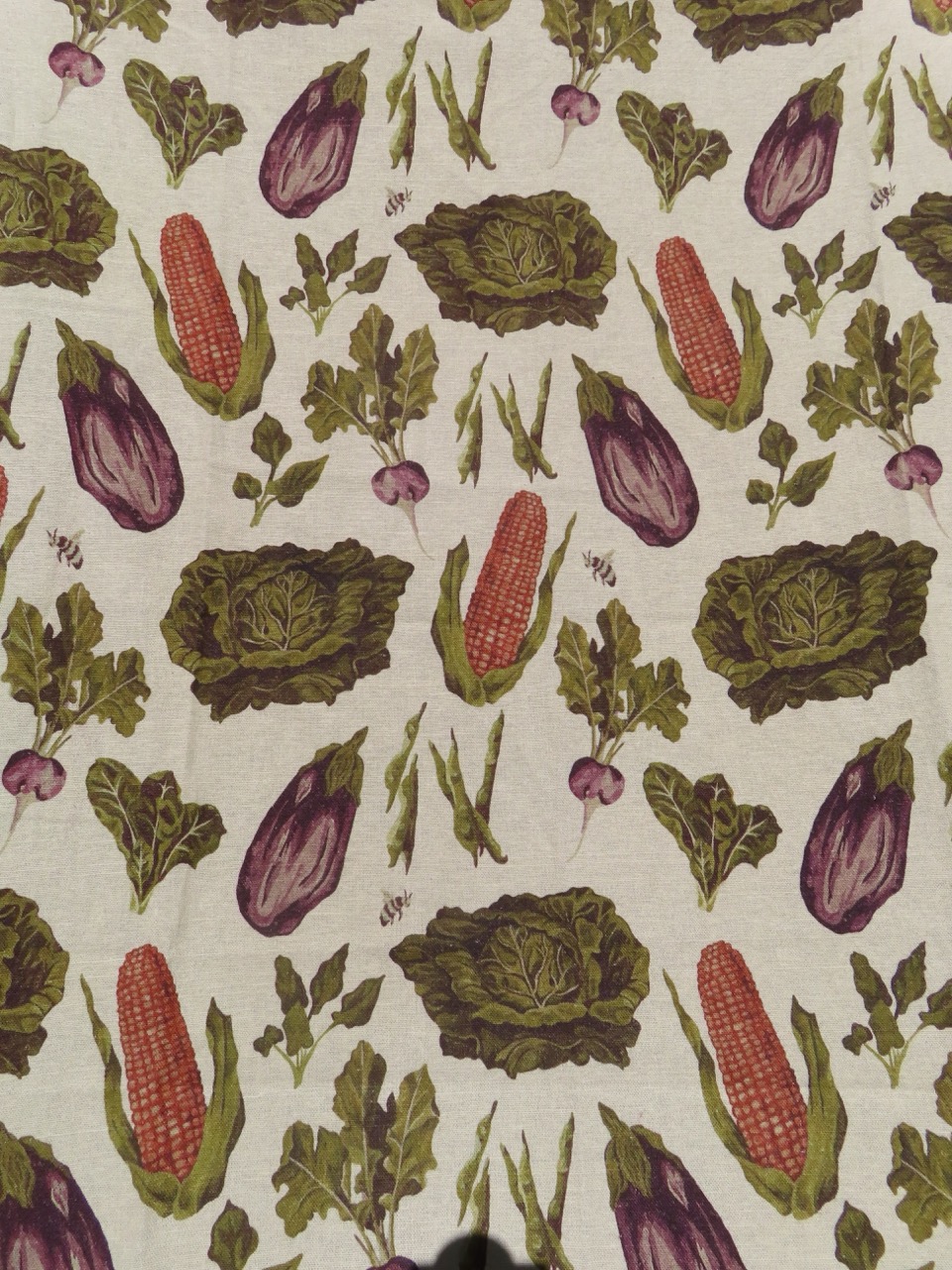 A patterned fabric showcasing various vegetables, including red corn, eggplants, lettuce, and beets, arranged in a symmetrical design on a light-coloured background.