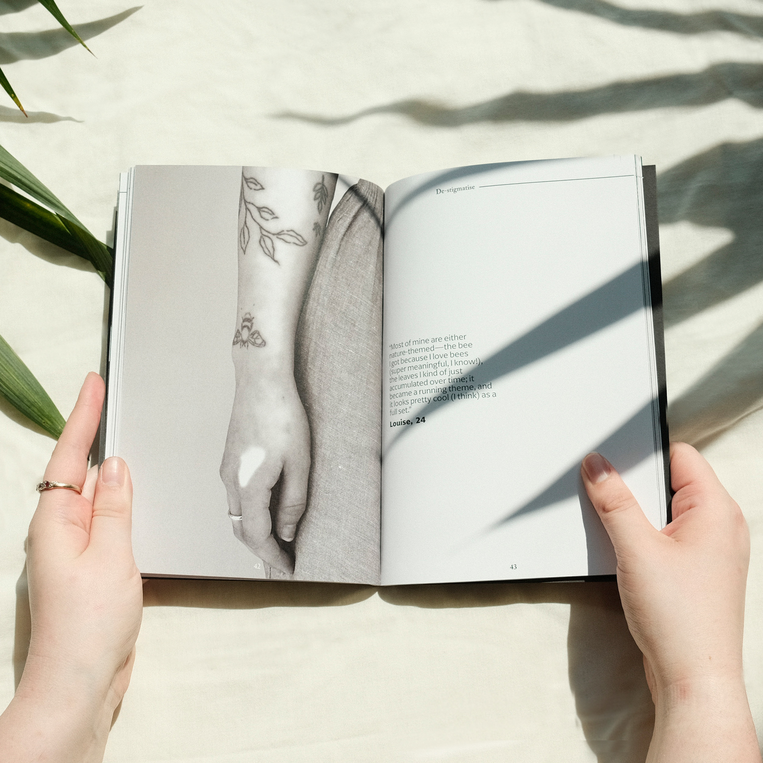 Hands holding open book showing black and white illustration of tattooed arm on left page, text on right page, with plant shadows