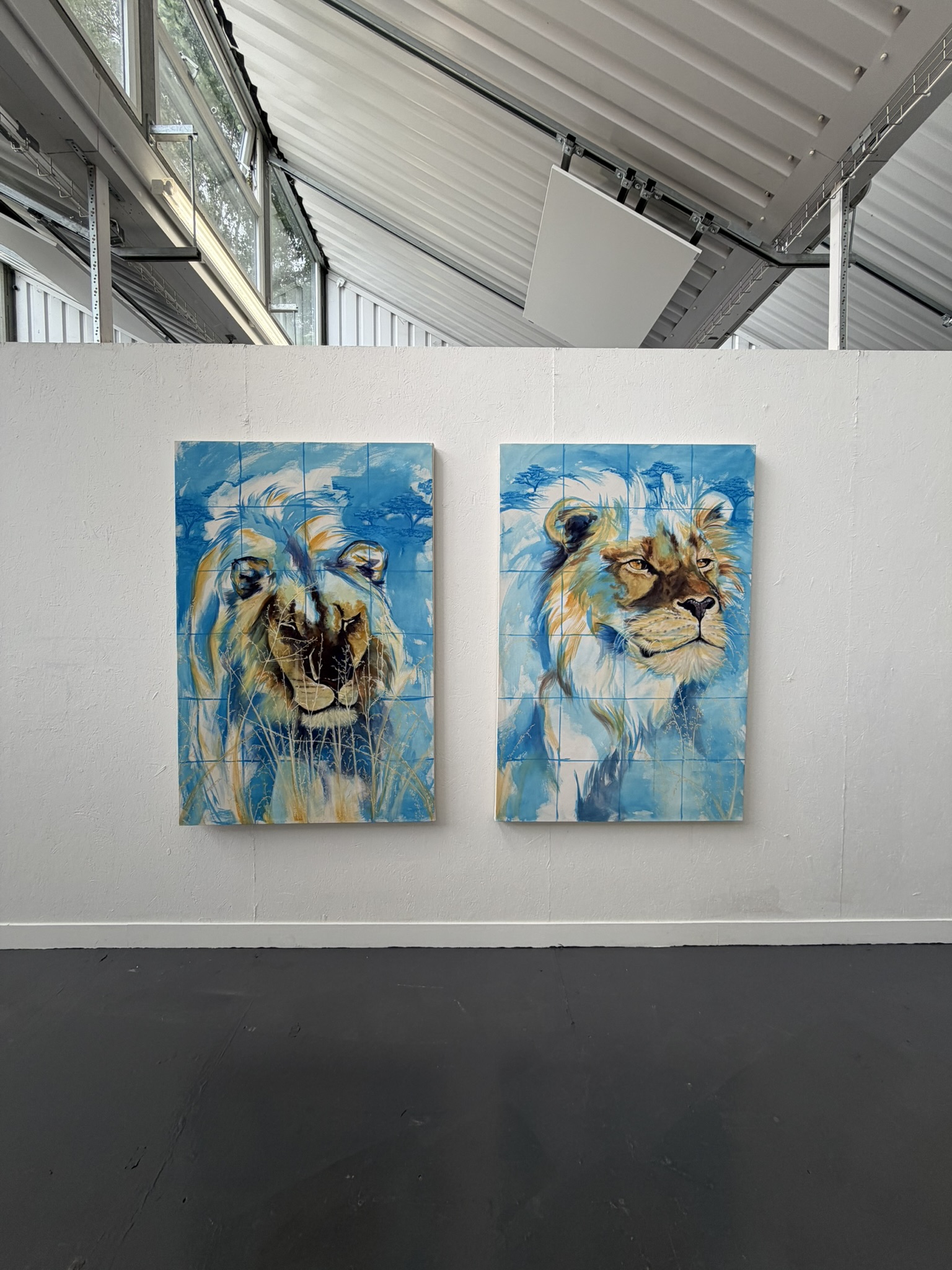 Two large paintings of lions displayed on a white wall. The left artwork shows a lion's face with a dark mane, while the right features a lion with lighter fur and a prominent mane. Both paintings use bright blues and yellows for the background.