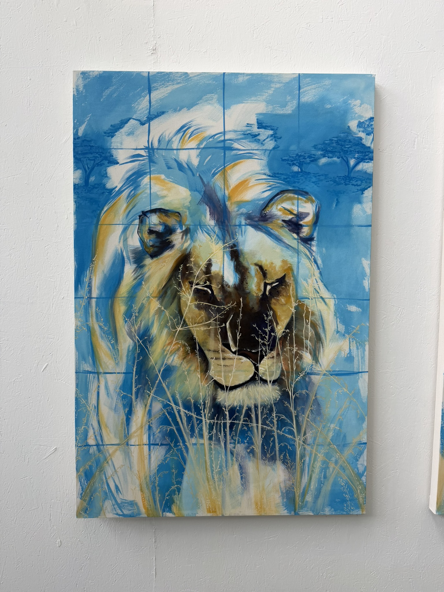 A large painted artwork of a lion's head displayed on a blue background. The painting features vibrant colours and textures, with hints of grass in the foreground, and is divided into square sections, enhancing the overall visual effect.