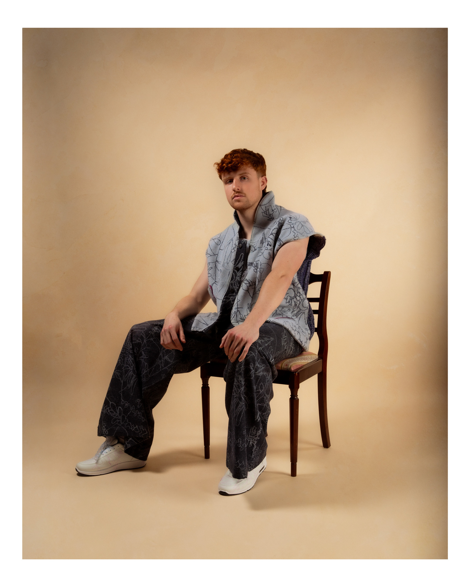 Model sat on a chair wearing baggy, patterned fit, beige background.
