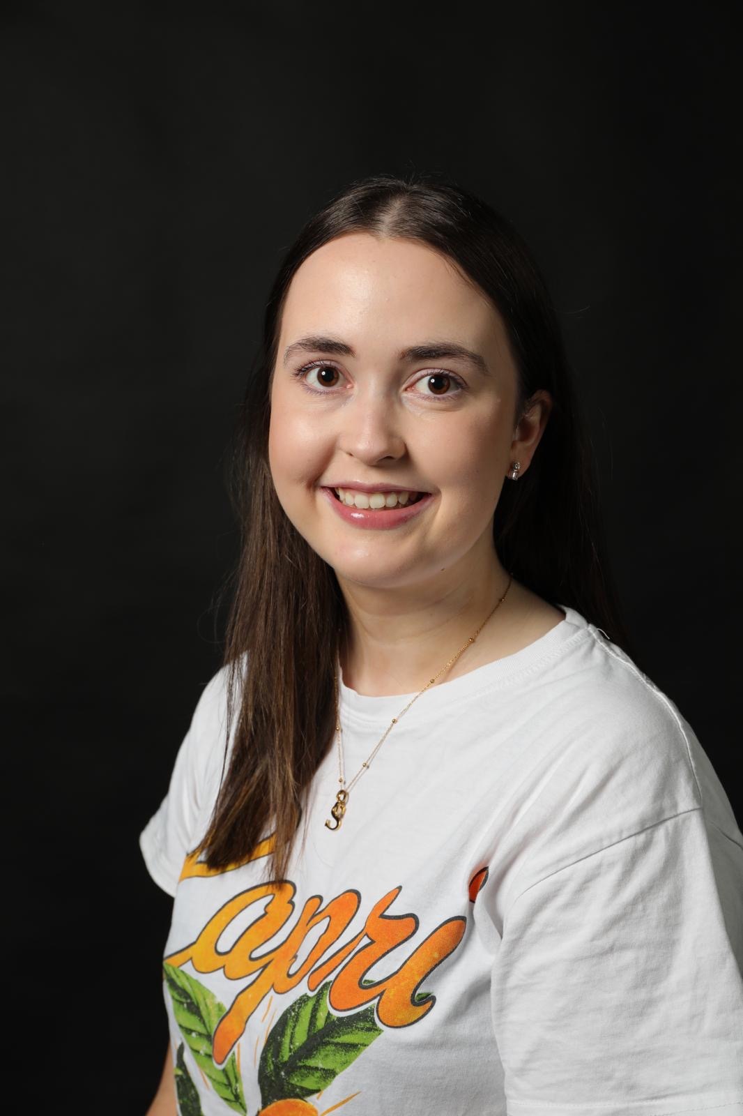 Young woman with long brown hair wearing white t-shirt with orange text and green leaf design, smiling against black background.