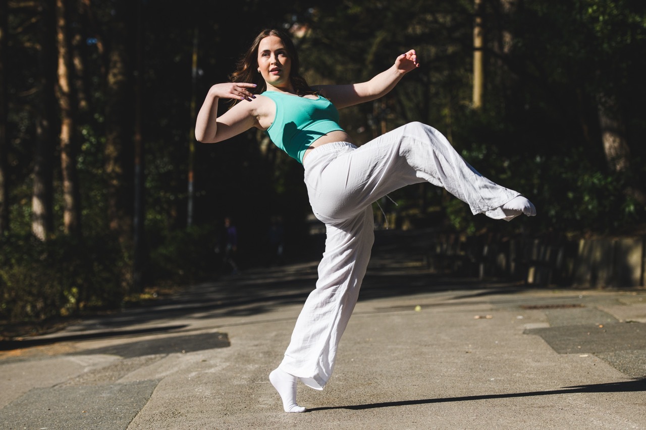 Woman in turquoise top and white trousers performing dance kick on outdoor paved surface with trees in background.