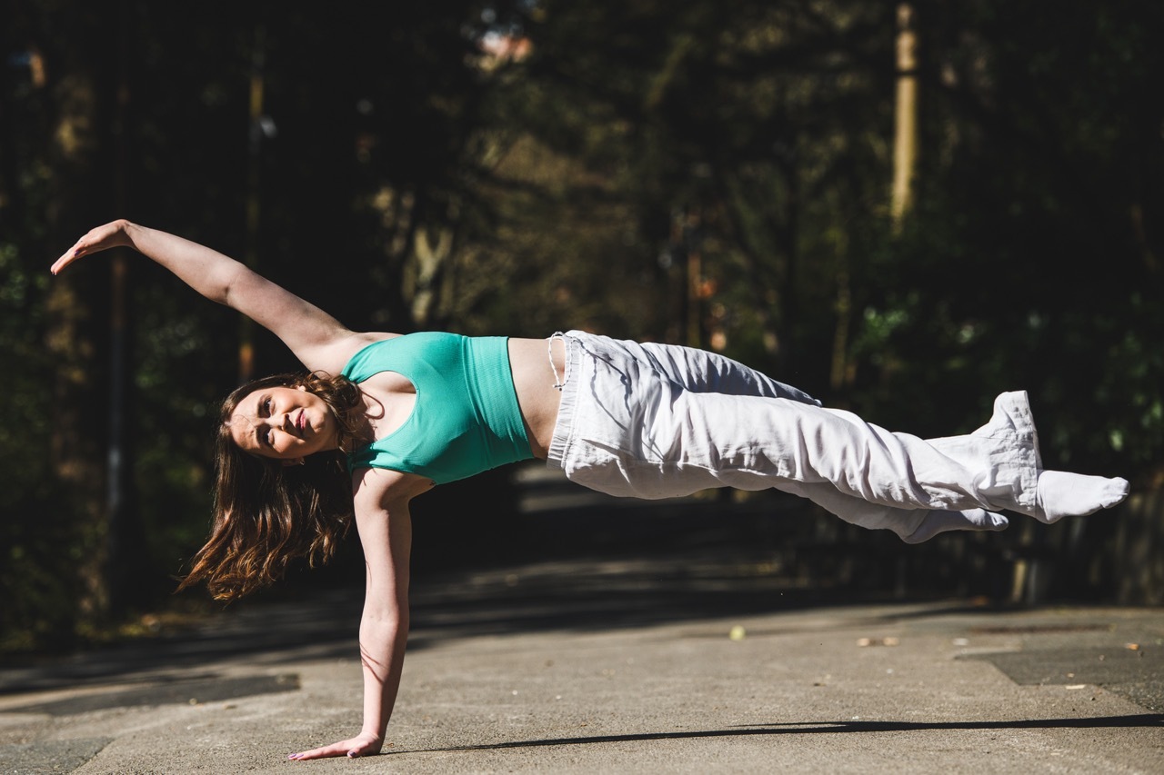 Woman in teal top and grey trousers performing side plank on outdoor pavement with trees in background.