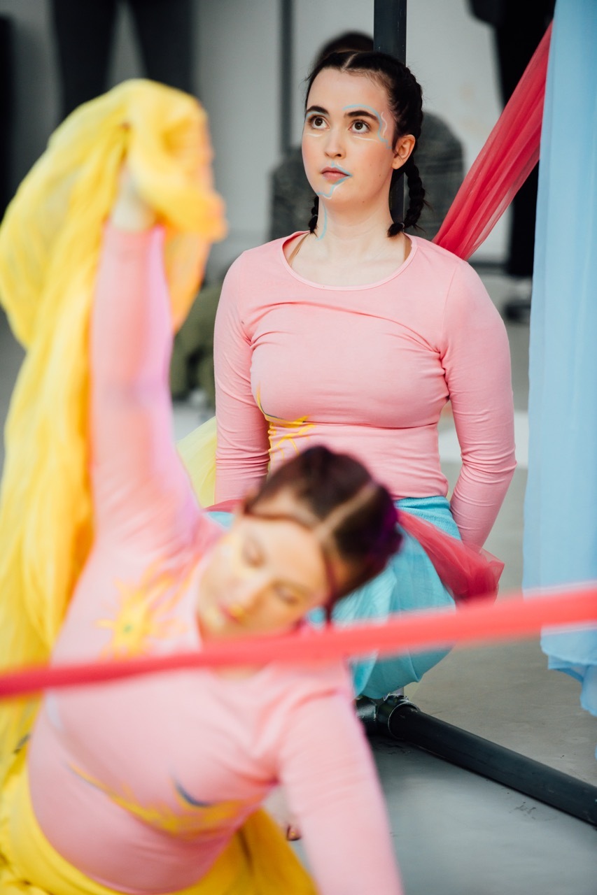 Dancers in pink costume wear amongst large translucent fabric pieces in yellow, pink, and red colours in bright indoor space.