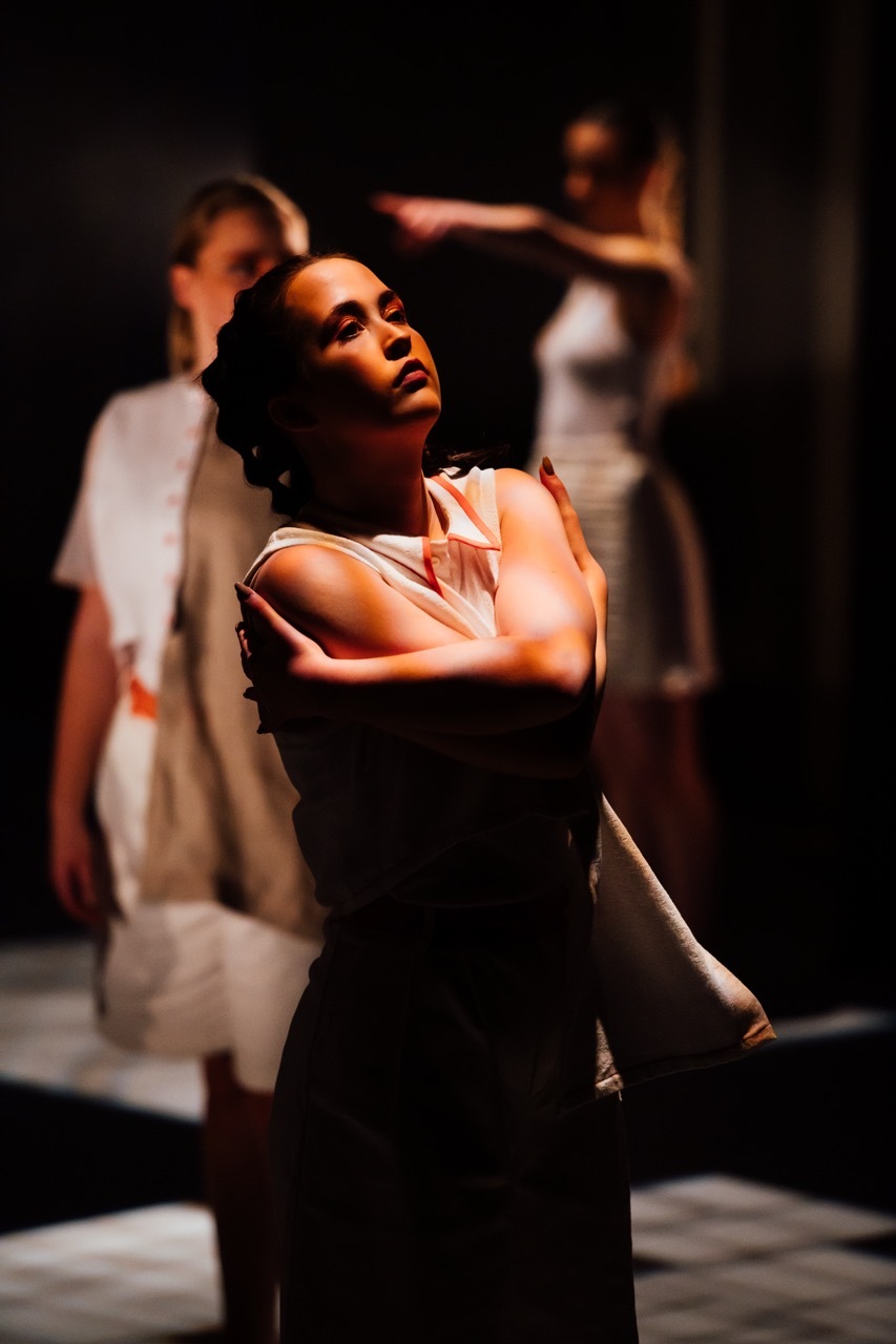 Woman in white top with arms crossed looking upward, two blurred figures in white clothing in background, dramatic lighting on stage.