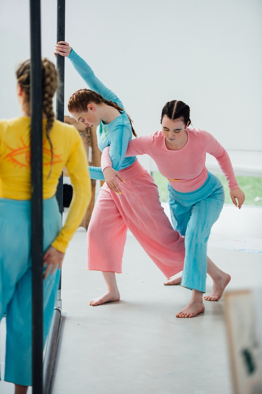Two women stretching at ballet barre wearing pink and blue dance costume, with mirror reflection visible on left side.