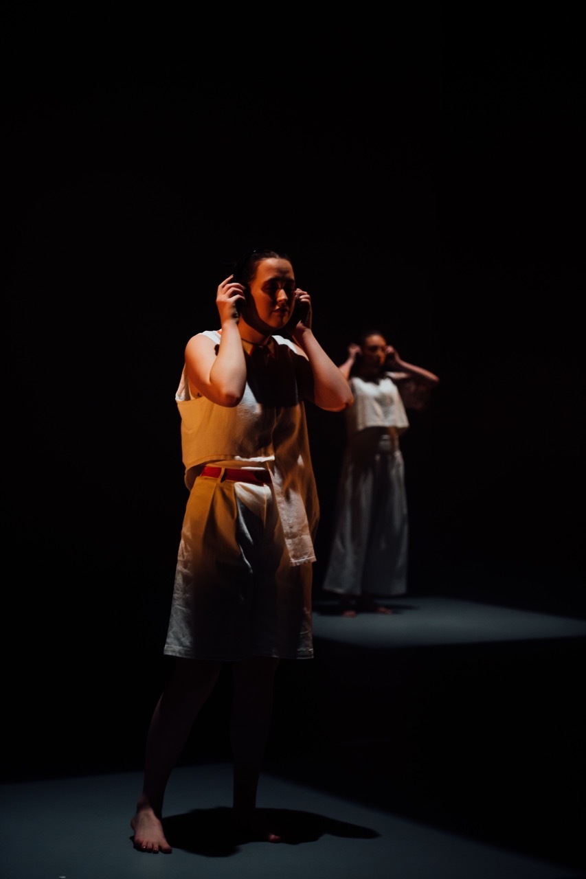 Performer in beige costume with hands raised to head on dimly orange-lit stage, additional figures visible in background.