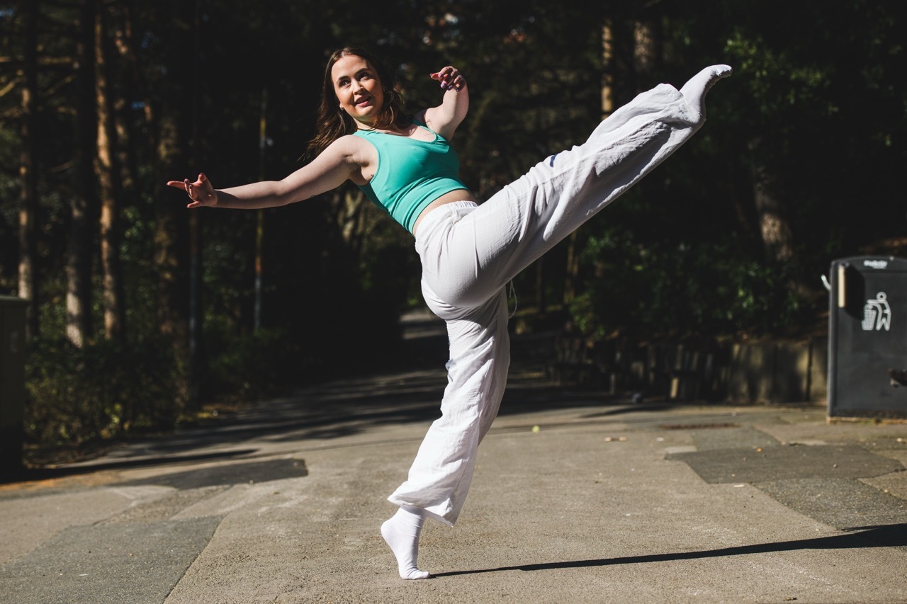 Woman in turquoise top and white trousers performing high kick on outdoor paved surface with trees in background.