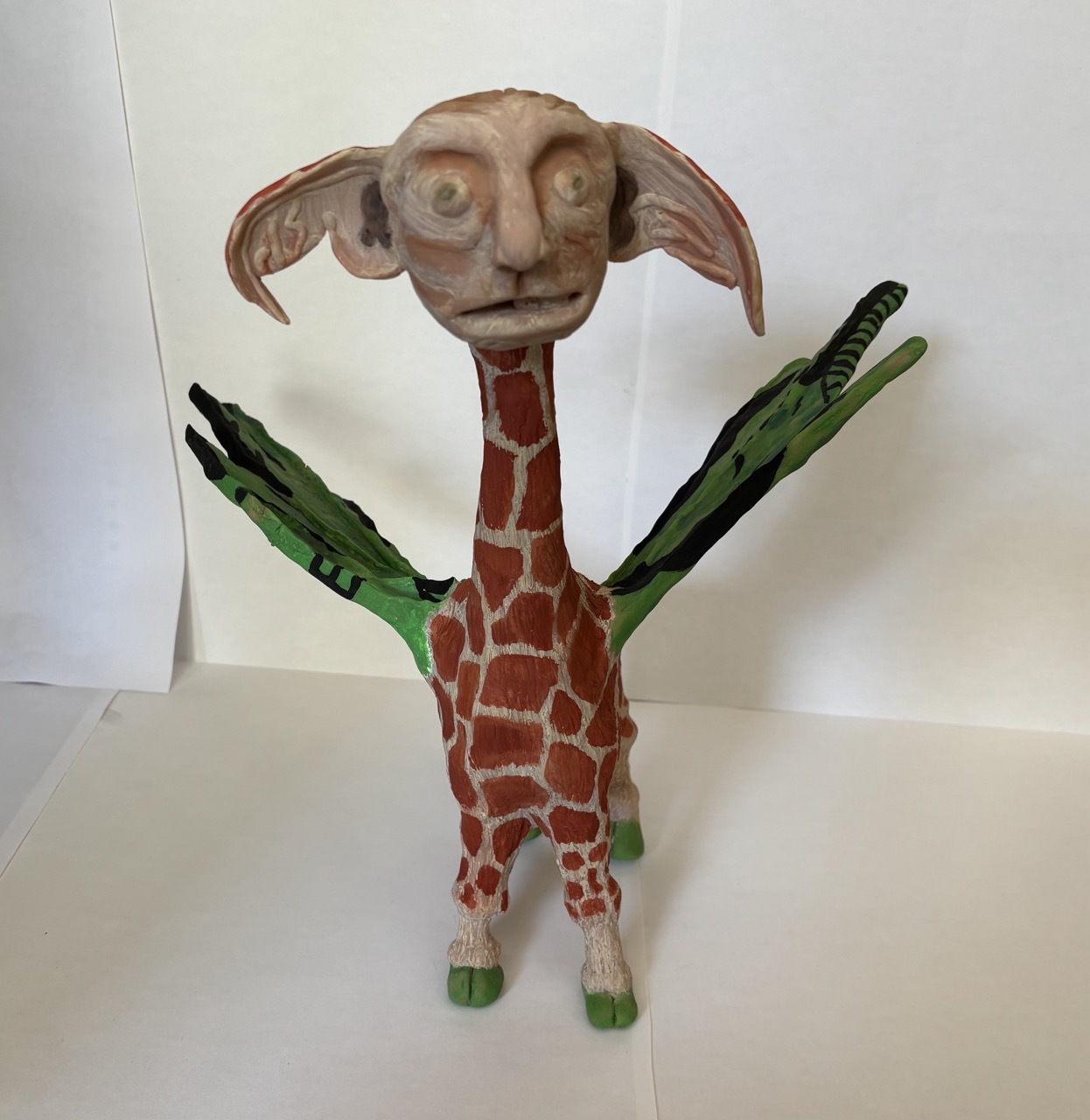 Clay sculpture of giraffe-like figure with large ears, brown giraffe-pattern body, green striped wings and green feet on white surface.