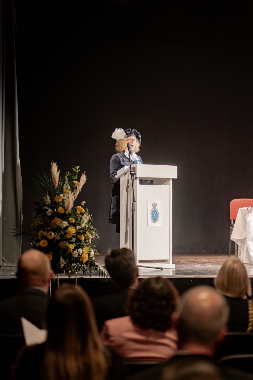 A woman in a hat speaks at a podium on a stage, with a large floral arrangement to the side and audience members seated in the foreground.