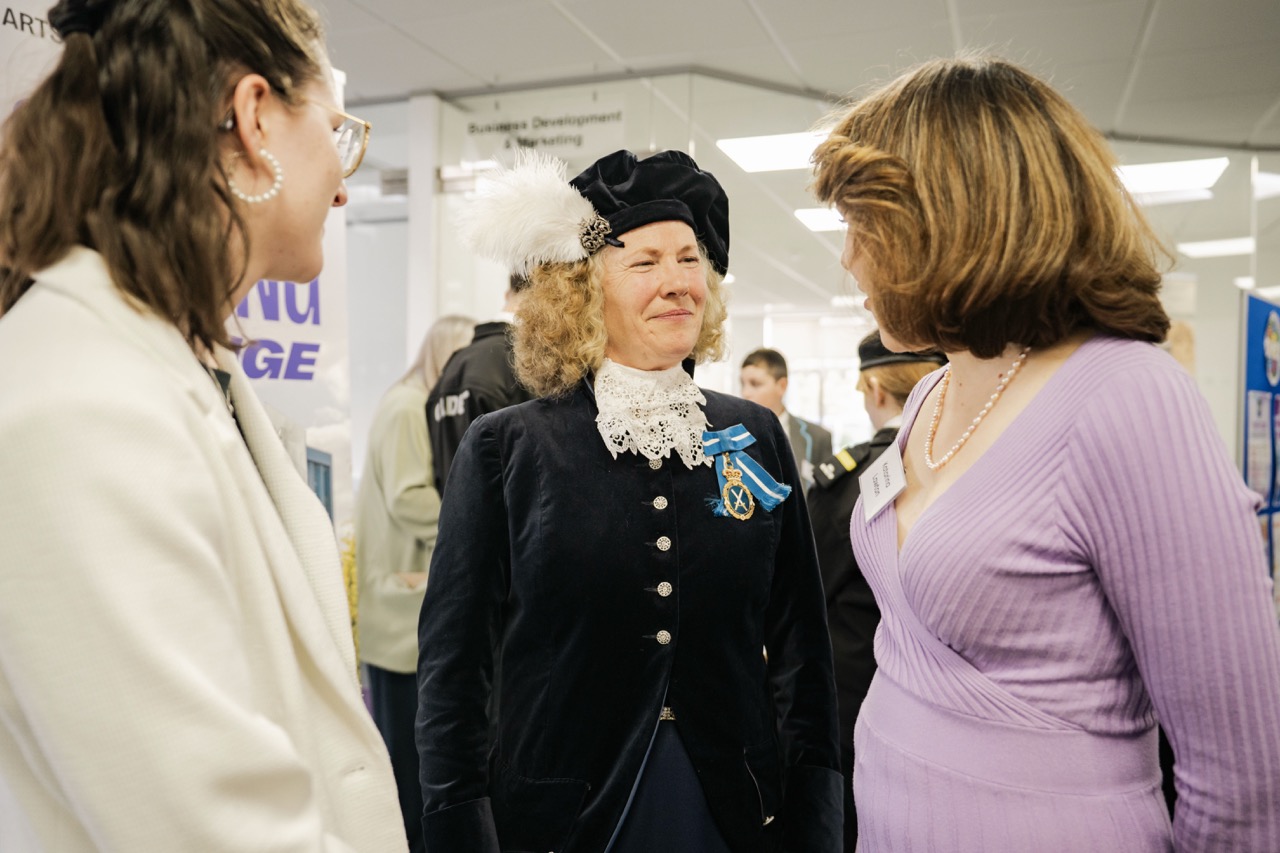 Three women stand together in an indoor exhibition space, with the central woman wearing a black historical-style outfit and hat, and display stands visible in the background.
