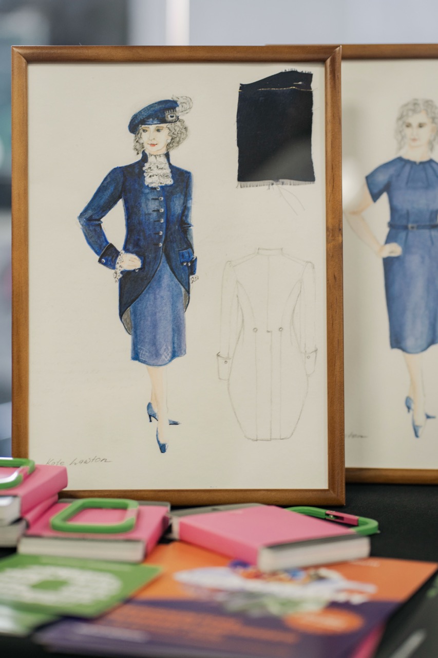 A framed fashion sketch of a woman in a blue suit and hat is displayed beside another sketch, with colourful notebooks and fabric swatches on a table in the foreground.