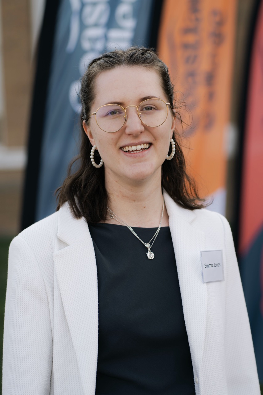 A woman wearing round glasses, pearl earrings and a white blazer stands outdoors, with blurred vertical banners in the background.