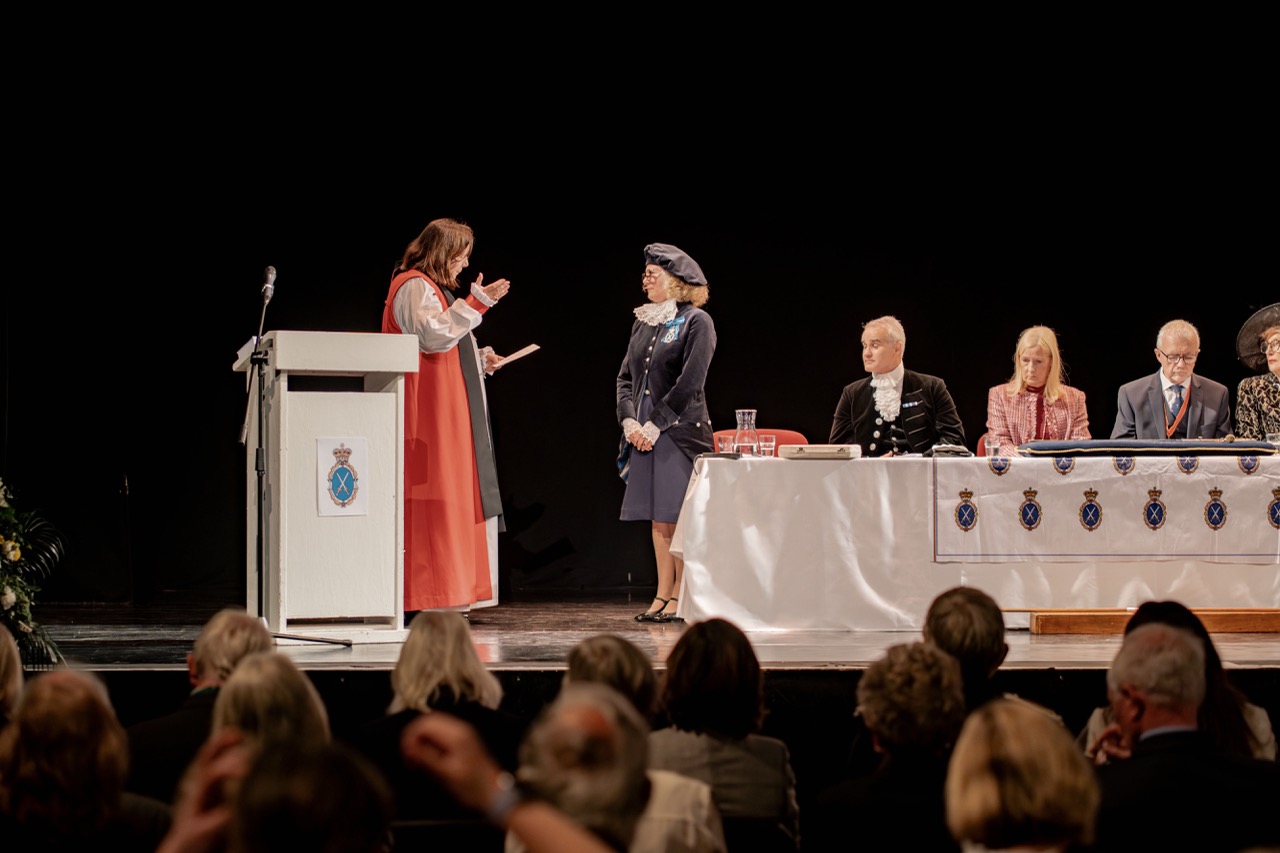 A woman in a red robe presents an award to a uniformed woman on a stage, with a panel of seated officials at a draped table and an audience in the foreground.