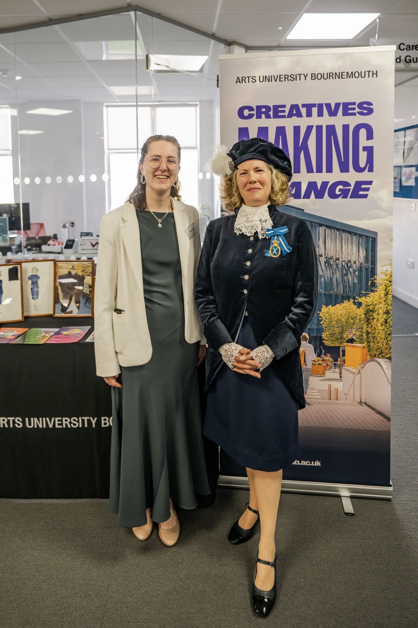Two women stand indoors beside an Arts University Bournemouth banner reading “Creatives Making Change”, with exhibition displays and a table of printed materials behind them.