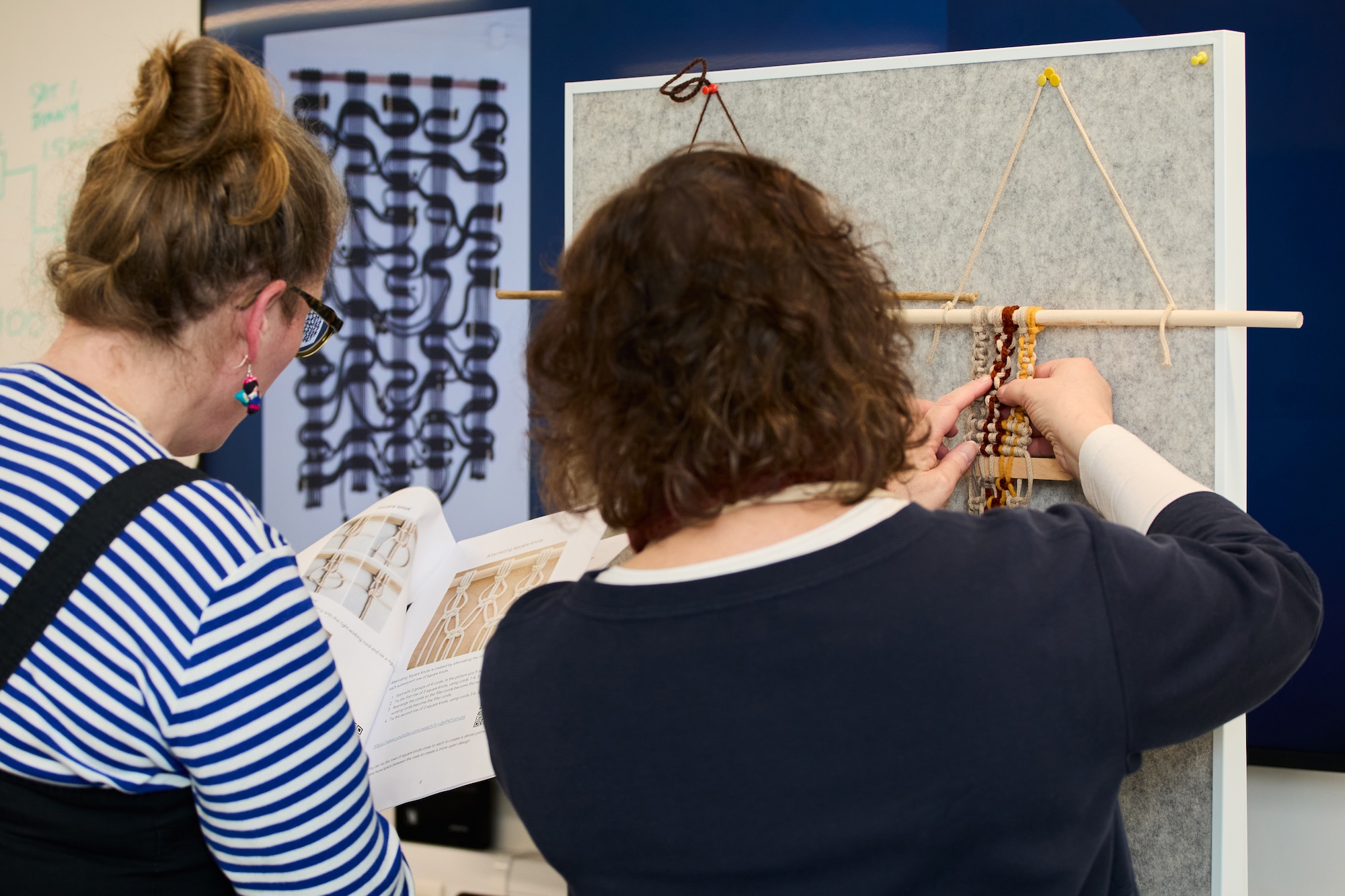 Two people examine a textile sample on a display board in a studio, with one holding an open book and patterned prints visible behind them.