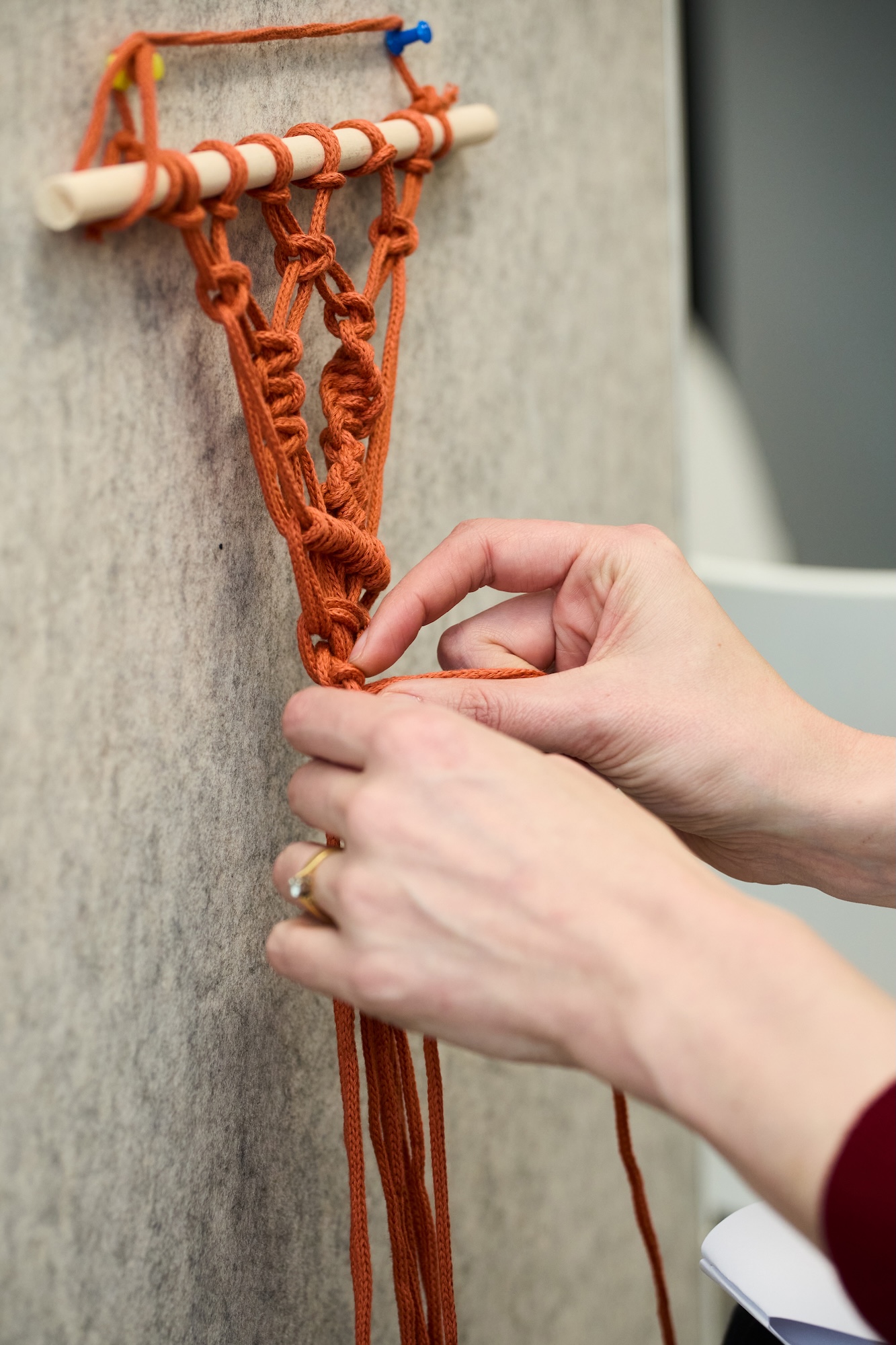 Hands weaving orange cord into a macramé wall hanging on a wooden dowel, with a grey textile background.