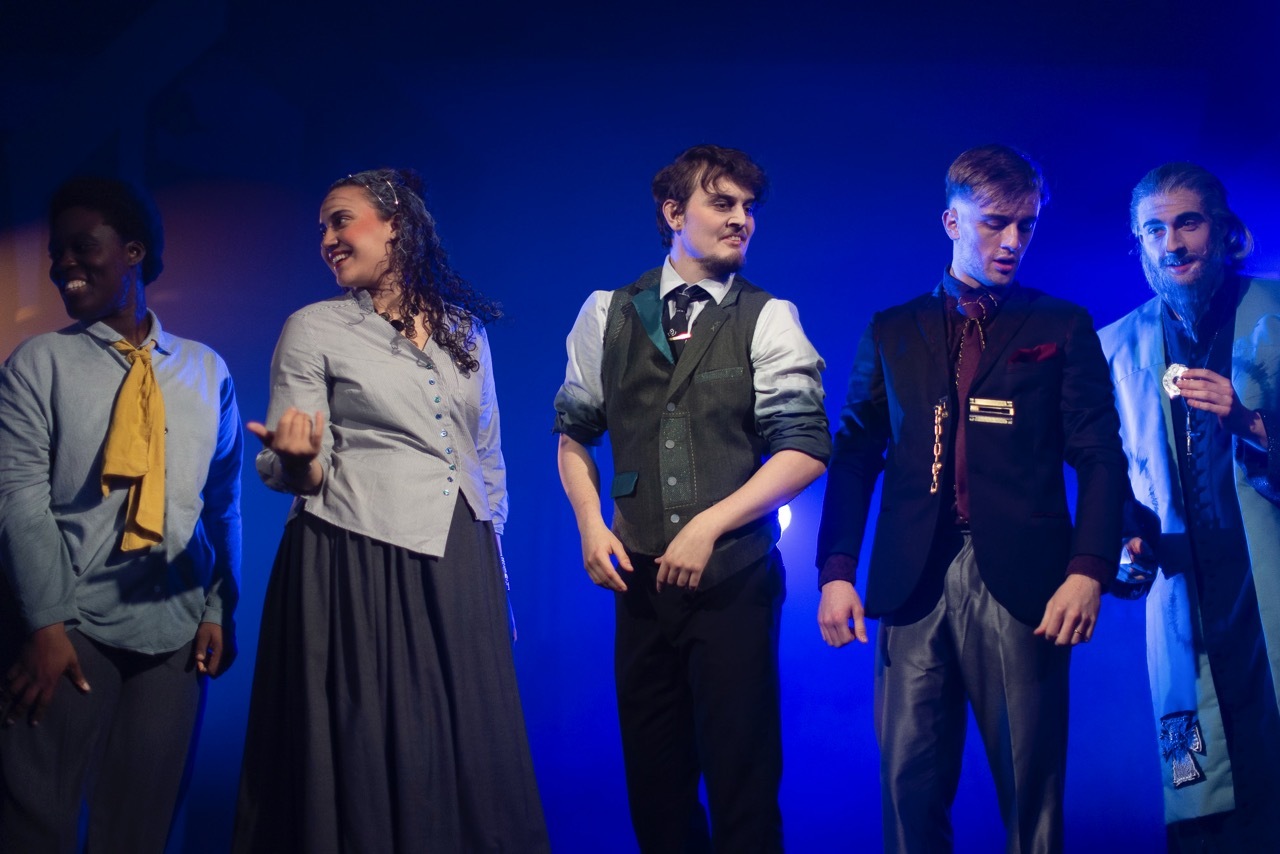 Six actors on stage in period costume under blue lighting, wearing grey and dark clothing including waistcoats and long skirts.